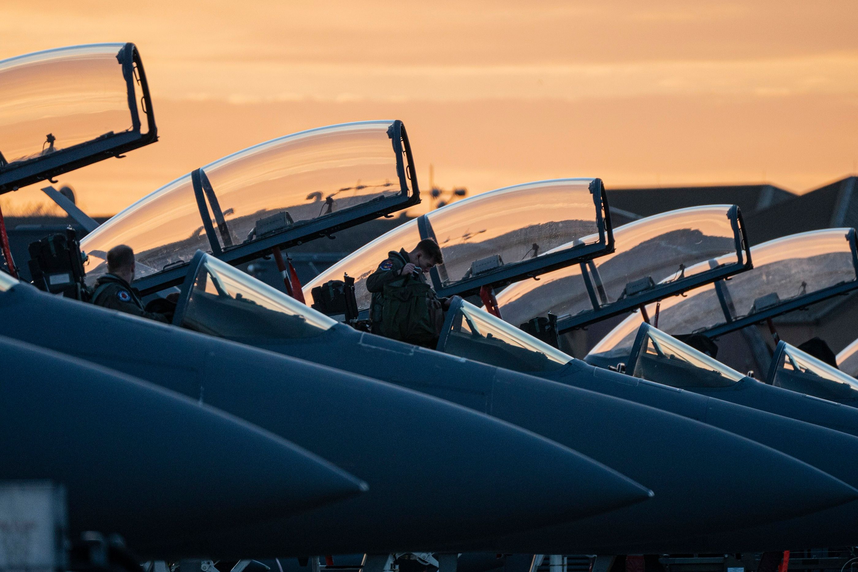 A row of F-15E Strike Eagles with canopies open against an orange sunset as aircrew climb in